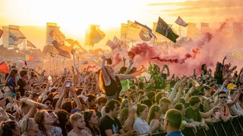 PA Wide view of the crowd at Glastonbury near sunset, with lots of flags and flare smoke visible. 