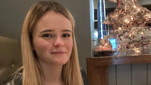 Family handout  Amelia Butlin with blonde hair, gazing at the camera with a small faux Christmas tree in the background. 