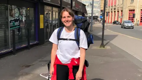 Chloe Davies Chloe Davies has her hair tied back and is wearing a white t-shirt and black bottoms, with a red jacket tied around her waist. She has a large blue and black backpack on her back. She is smiling at the camera as she stands on the pavement next to an urban road