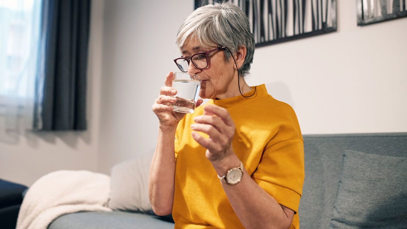 A female wearing glasses taking a supplement with a glass of water.