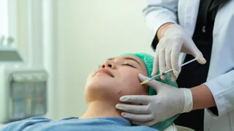 Getty Images A woman in a blue medical gown and green medical gap lies down with her eyes closed, as a doctor in a white coat and gloves injects a syringe into the woman's face