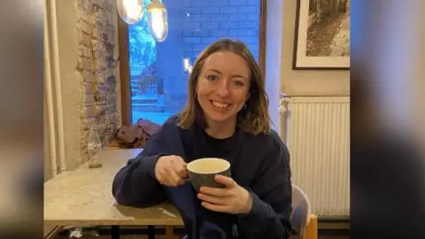 Gemma Byrne/Mind Gemma sits at a table holding a mug and smiling, she is wearing dark clothes.