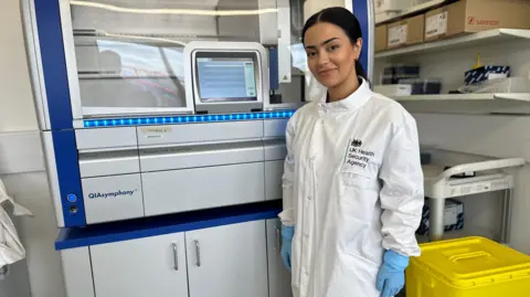 A woman with brown hair in a ponytail smiles at the camera wearing a white lab coat and blue rubber gloves. She's standing in front of a machine for processing biological samples in a lab