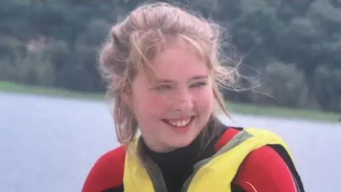 Marina Waldron in a red and black wetsuit and a yellow and grey lifejacket with open water behind her.