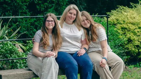 Robert Timothy/BBC Charlene sitting on a parkbench with her two daughters, Isla and Skye