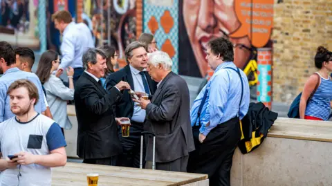 Getty Images Group of many businessmen people standing outside in England at the Shipwrights Arms pub tavern bar drinking beer from cups