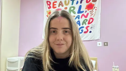 Rachel/BBC Rachel, who has blonde hair and is wearing a black jumper, sits on a bed with a set of portable draws beside it. A white banner on a purple wall behind her is partly obscured by her head, but the colourful letters on it appear to include the words 'Beautiful girl'