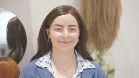 A picture of Megan McCready, who has alopecia, smiling. She is wearing a wig and is pictured in front of several other wigs.