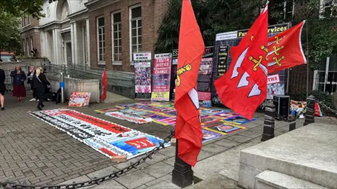 Richard Knights/BBC Placards and posters outside the Civic Centre in Chelmsford. They are mostly accusing the Essex Partnership University NHS Foundation Trust of causing the deaths of people. They are black with colourful font.