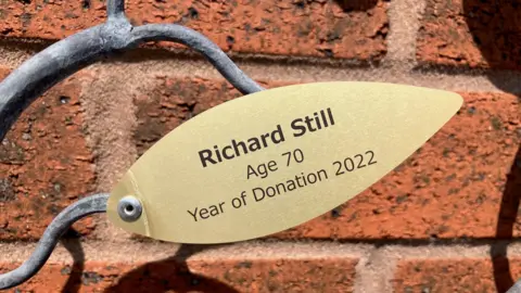 A gold-coloured piece of metal in the shape of a leaf is attached to a wavy piece of grey-coloured metal, with a redbrick wall behind it. The inscription in black writing on the leaf says: 