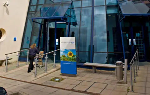 Alamy An outdoor shot of the entrance of the South West London Elective Orthopaedic Centre, a glass building with an NHS sign at the front, with nn employee walking up to the door pushing a trolley, taken in Epsom in 2009