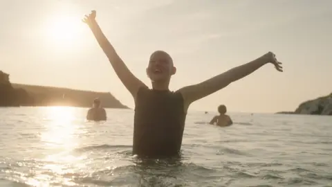 BBC/October Films/Neil Bonner Kris is standing in the sea. She has her arms outstretched and is smiling. Two people can be seen in the background behind her and the sun is shining.