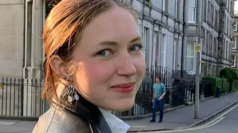 Gareth Williams Rhiannon has brown, tied back hair and is wearing a black leather coat and white blouse. She is turned to the camera and is smiling. Behind her are a row of houses in Edinburgh where she was studying at university.