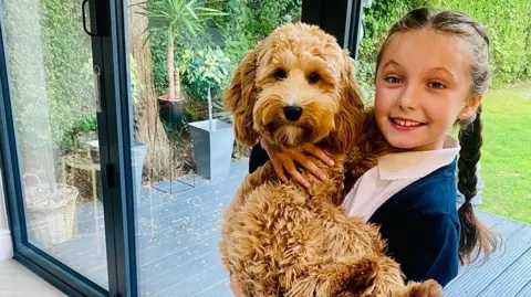 Family handout A little girl in school uniform with two plaits holding a puppy. 