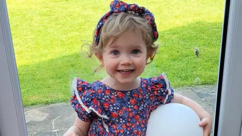 Family photo Isabelle Cooper holding a balloon at the door into the garden. She is wearing a blue and red floral dress with matching headband. 