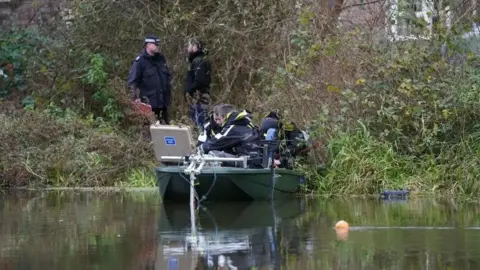 PA Media Divers and police officers in a small boat filled with equipment near a bank on the River Wensum. Two men are standing on the bank, with a building visible behind trees and bushes.