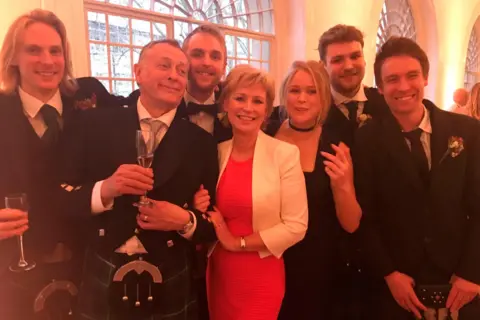 Sally Magnusson  Sally Magnusson with her family in formal wedding clothes. Sally is in the centre wearing a red dress and white jacket and her daughter Anna Lisa is on her right wearing a black dress. Sally's husband Norman is on her left in a kilt and sporran and formal wedding jacket. Also pictured are the couple's four sons Jamie, Rossie, Magnus & Siggy.