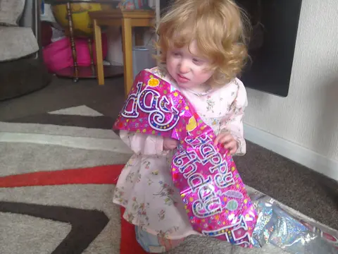 Lisa Hawker Young Jadyi with curly hair and a pink floral dress holding a vibrant pink banner than says Happy Birthday