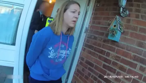 Cheshire Constabulary A still from police body-worn camera footage showing the moment of Lucy Letby's arrest, as she leaves a house with a birdbox on the wall, her hands cuffed behind her back, wearing a blue Lee Cooper hoodie and with a stricken expression on her face