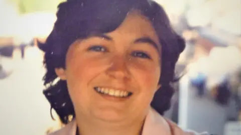 Family A woman with mid length brown hair smiles towards the camera, the collar of the pink top or coat she is wearing is barely visible. The background is out of focus