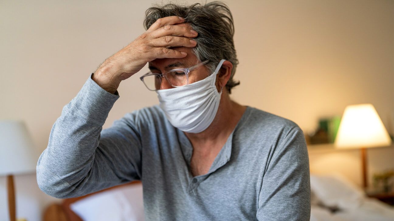 A sick older man wearing a face mask massages his head inside a room