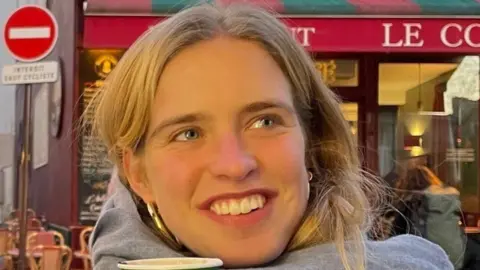 Hilary Nicholls Clarissa Nicholls, who has light hair and is wearing a grey top, smiling while outside a coffee shop in Paris