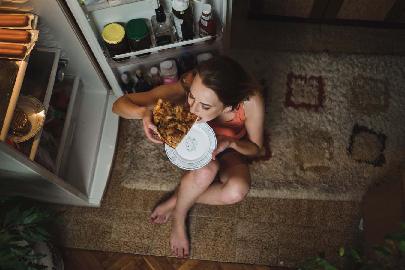 Woman sitting in front of the refrigerator and eating pizza slice