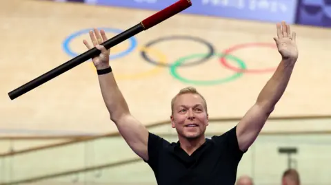 Getty Images Chris Hoy holds a baton in the air during a ceremony at a velodrome in Paris. Behind him is the velodrome with the five Olympic rings