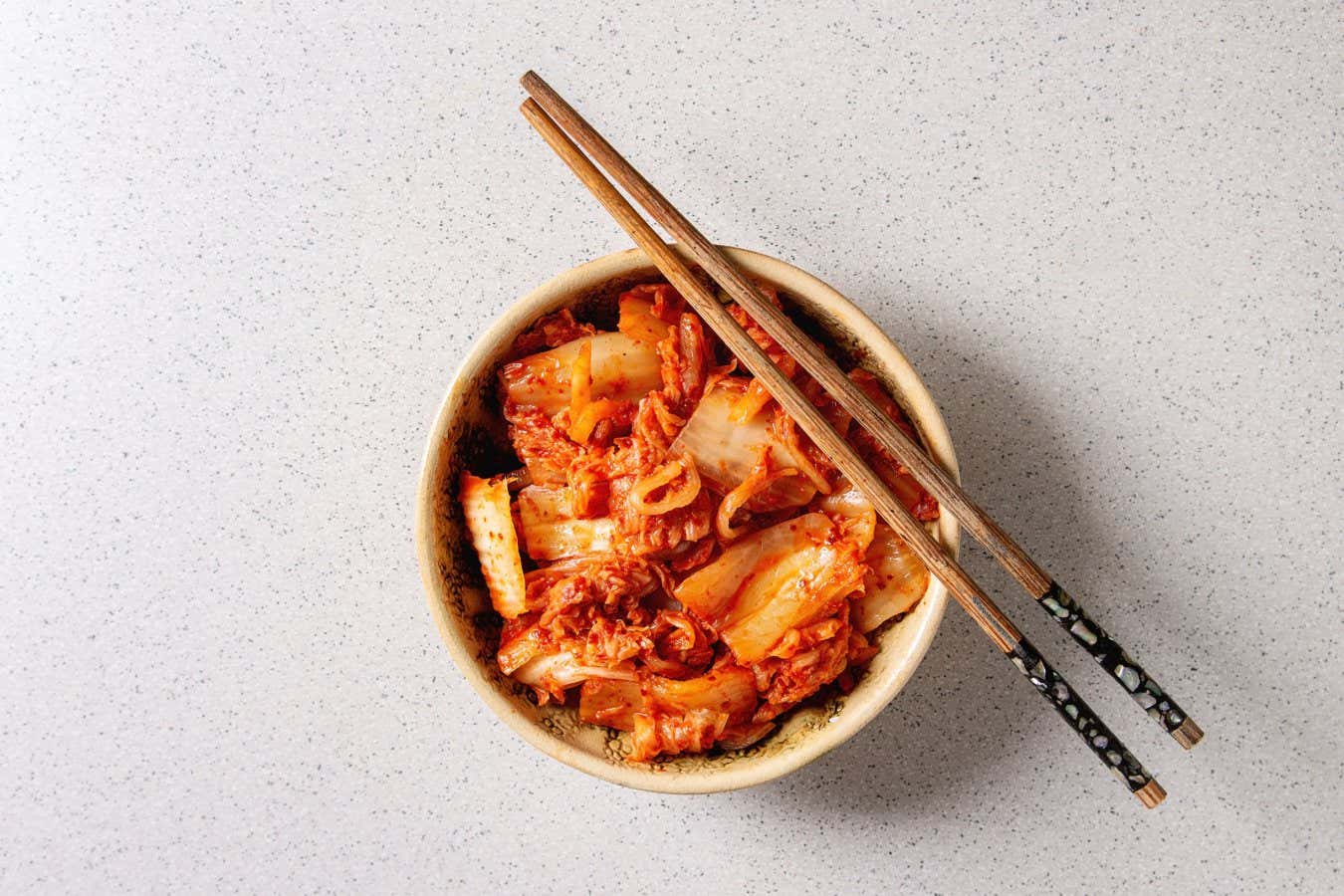 Korean traditional fermented appetizer kimchi cabbage salad in ceramic bowl with shopsticks over grey spotted background. Flat lay, space.; Shutterstock ID 1343369669; purchase_order: -; job: -; client: -; other: -