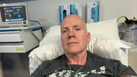 Larry Lowe Larry in a grey t-shirt laying down on a hospital bed next to a heart monitor. He is looking directly at the camera 