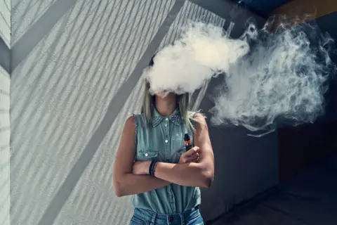 Getty Images Young lady wearing blue demin shirt and jeans, She has her arms crossed and is holding a disposable vape, with a plume of smoke obscuring her face. 