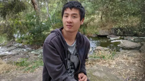 Supplied Tim Chan is sitting next to a stream in an outdoor area. There are trees visible behind him, and he is wearing a dark grey hooded sweatshirt and looking straight at the camera. 