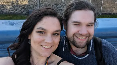 HANDOUT Kari, who has long brown hair which is tied back and is wearing a blue knitted jumper, sits close to his partner Kaitlyn, who has long brown curled hair and wears a strappy black top, as they smile for a selfie outside the Tower of London.