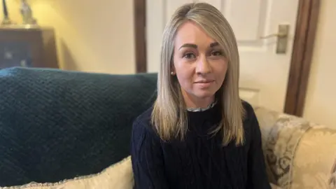 A woman sitting on a sofa looking at the camera. She has straight blonde hair and is wearing pearl earrings and a dark blue cable knit jumper. The sofa is cream coloured with a dark blue throw over the back and there is a door in the background. 