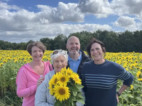 Glenn standing in front of a field of sunflowers with, from left to right, his sister Lorna, his mum Jennifer and his wife Claire. They are all smiling and Jennifer is holding a bunch of sunflowers.