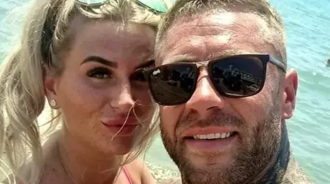 Family Handout A blonde-haired woman, Aaron Morris' wife Samantha, with sunglasses on her head and Mr Morris, who is smiling and wearing sunglasses. They are looking at the camera in front of the ocean, heads close together and taking a selfie. 