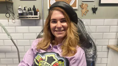 Alice Harding sits on a black chair covered in cling film smiling at the camera, she has light brown hair and a nose ring and is wearing a black beret