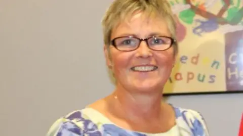 Betsi Cadwaladr University Health Board Sue Hill looking at the camera and smiling. She is wearing a beige dress with purple flowers. She has brown glasses on. It is a head and shoulders shot of her. 