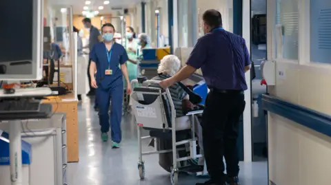 PA An NHS hsopital worker, wearing a surgical mask and scrubs, walks down a busy hospital corridoor 