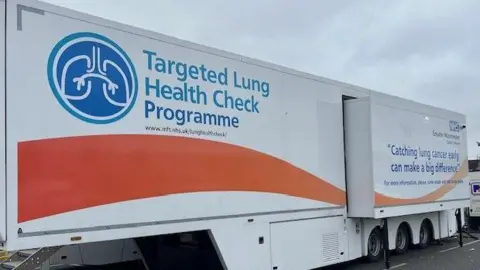 BBC A large white and orange lorry trailer with the words 'Targeted Lung Health Check Programme' painted in blue on the side, with an illustration of a pair of lungs next to it, parked up in a Tesco's car park in Greater Manchester.