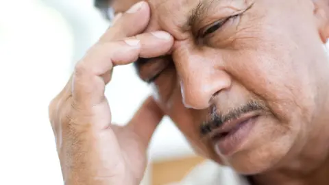 Getty Images Close-up of a man with dark hair and a moustache holding his right hand to his temple and grimacing in pain.