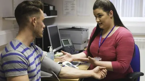 Getty Images A patient, wearing a blood pressure monitor on arm, getting assessed by a medical professional