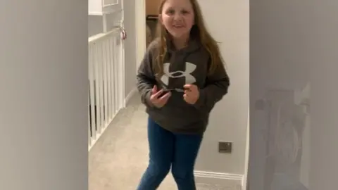 Claire Osborne A young girl standing inside her house in a sweatshirt and jeans, showing that both her legs bend inwards at the knees. She is smiling at the camera.