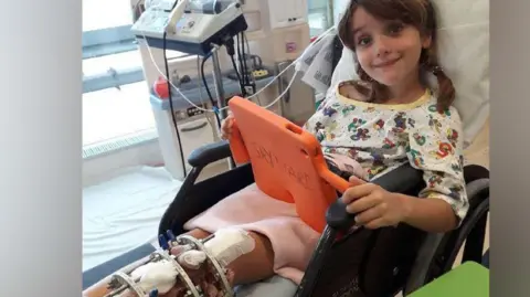 Dean Staltham A young girl lying in a hospital bed wearing pyjamas and looking at an ipad with a metal frame on her left leg.