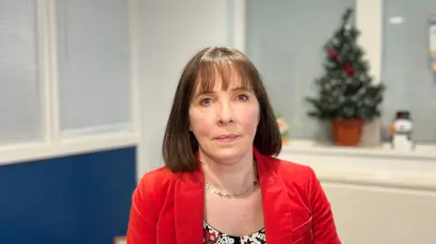 Joanne McClean. She has short brown hair with a fringe. She is wearing a red blazer, floral top and a necklace. 