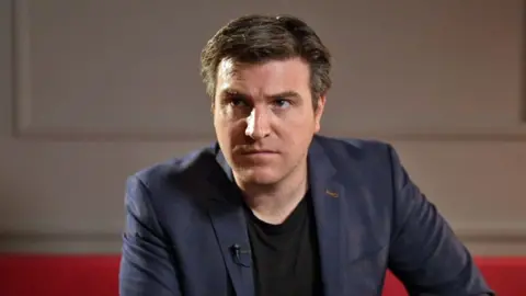 Headshot of Dr David Grimes who has short dark hair and is wearing a black t shirt and navy suit jacket. He is sitting on a red sofa with white wood panelled walls behind him. 