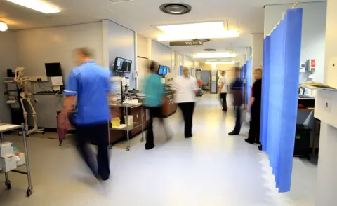 PA Media A busy hospital corridor with medical staff in motion, creating a blurred effect. Staff members in blue uniforms can be seen walking, with medical equipment and computer screens visible along the walls. The corridor has fluorescent lighting and a clinical appearance with white walls and blue accents.