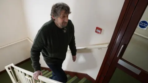 DAWID WOJTOWICZ/BBC Daniel Evans-Smith walking up the stairs to his flat in Northampton. He is holding onto a cream banister and is wearing a green wool jumper and navy cordroy trousers. His beard his slightly plaited and his hair is greying.