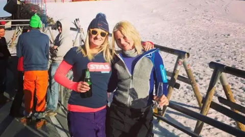 Susannah Morgan Susannah is wearing a wooly hat with a pom pom and holding a beer. She has her arm around a friend who has blonde hair and they are standing on the edge of a ski slope.