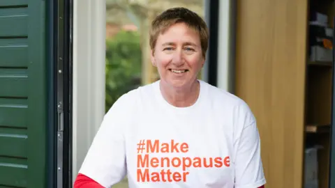 Jenny Smith Photography Diane Danzebrink in a doorway smiling at the camera. She wears a white T-shirt with the words '#Make Menopause Matter' written in orange letters in the centre. 